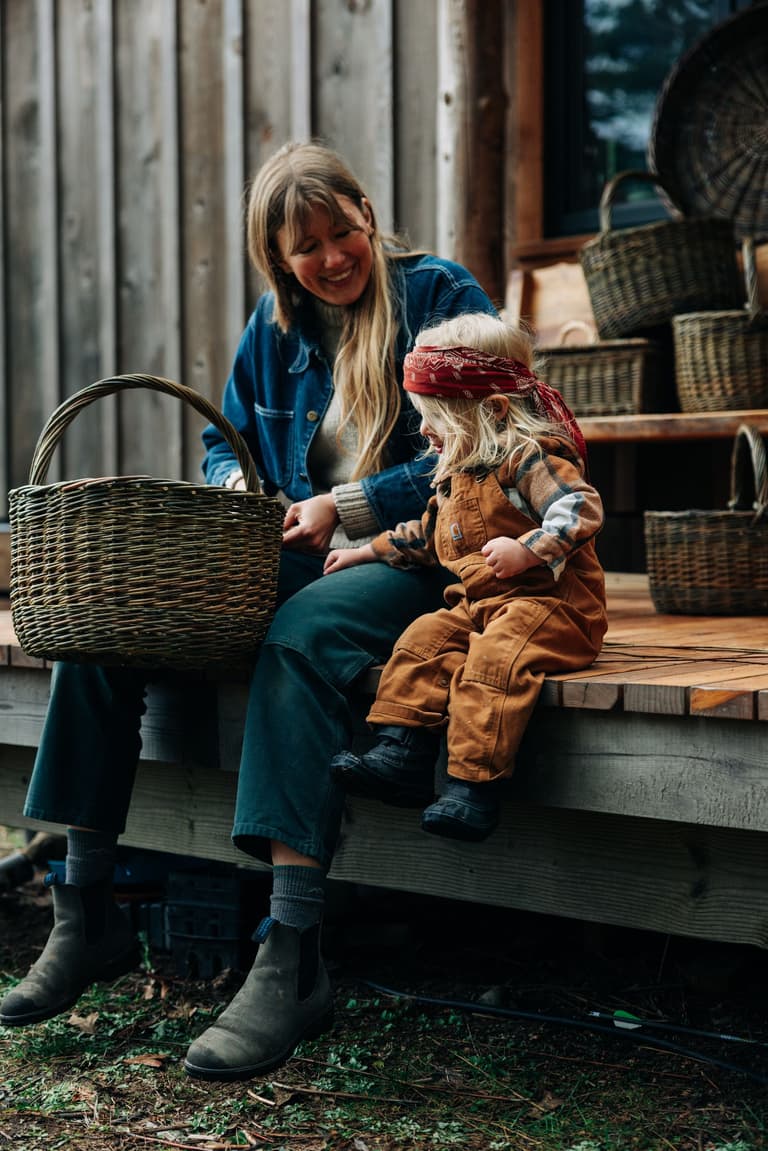Woman in denim shirt smiles while interacting with young child wearing red headband and tan jacket outdoors