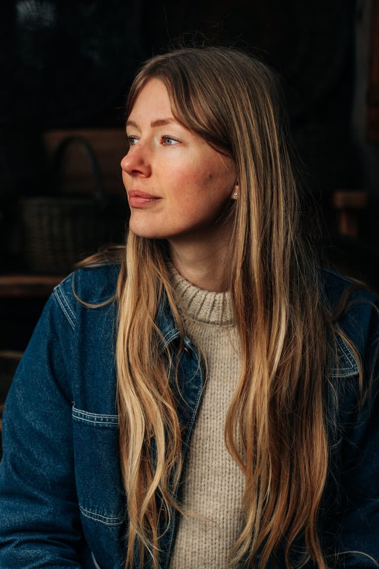 Alex Rooted in profile, wearing a cream sweater and denim jacket against a dark background