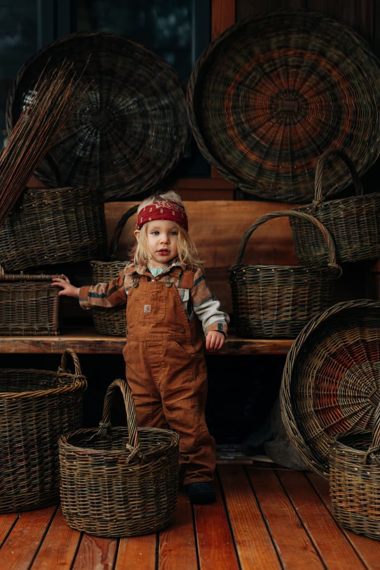Young child in rust-colored overalls and red bandana stands beside woven willow baskets in a wooden workshop interior