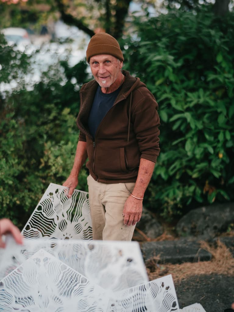 Smiling man in brown hoodie and tan beanie against blurred green foliage, outdoor portrait with natural daylight