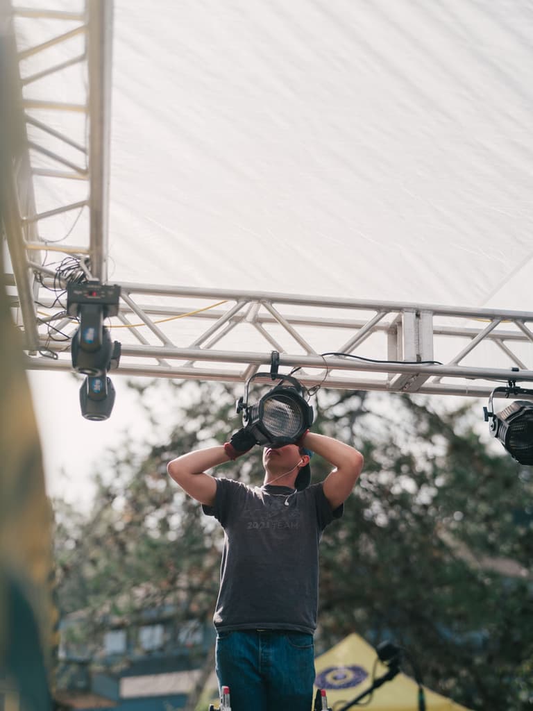 Technician holds vintage Fresnel spotlight against metal truss framework beneath overcast sky at festival setup