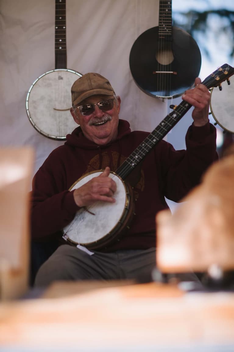 A smiling man in a ball cap playing the banjo, with more banjos hanging behind him