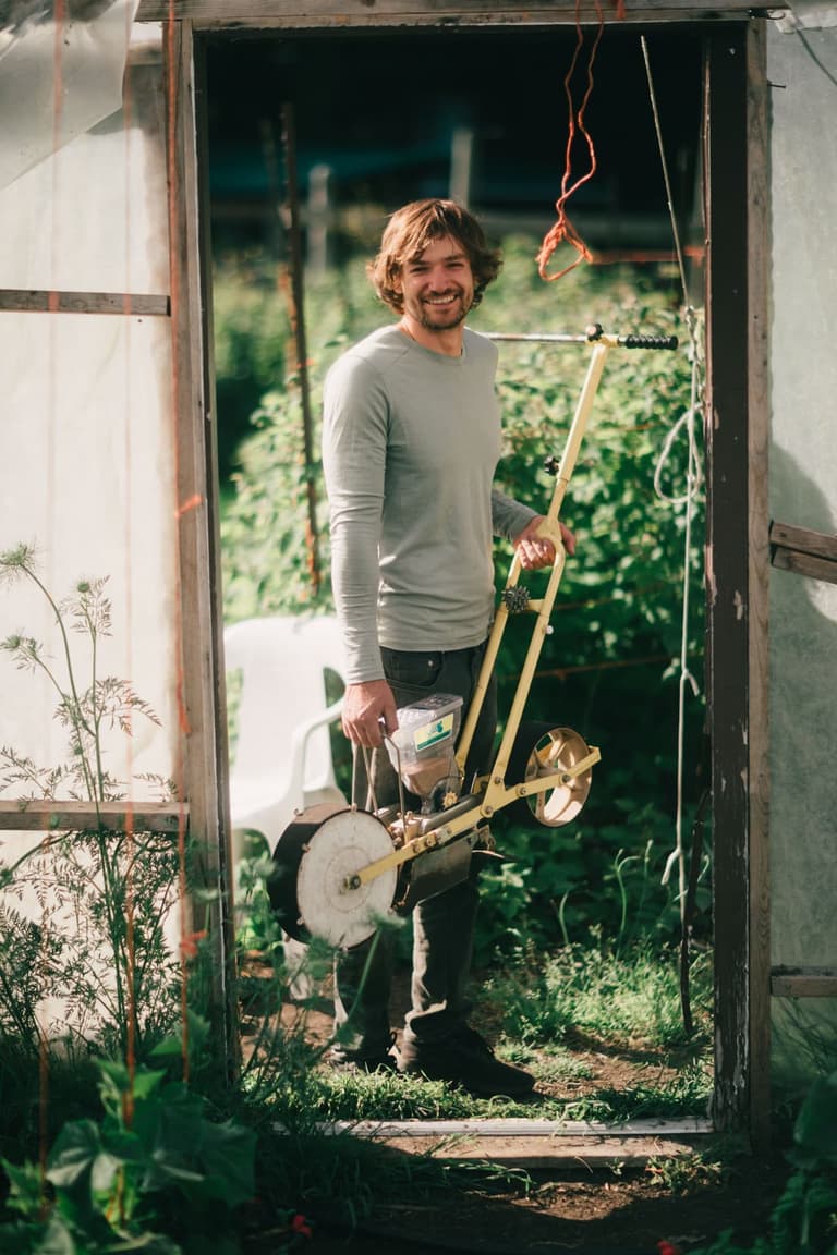 A grower standing in a greenhouse doorway holding a wheel hoe