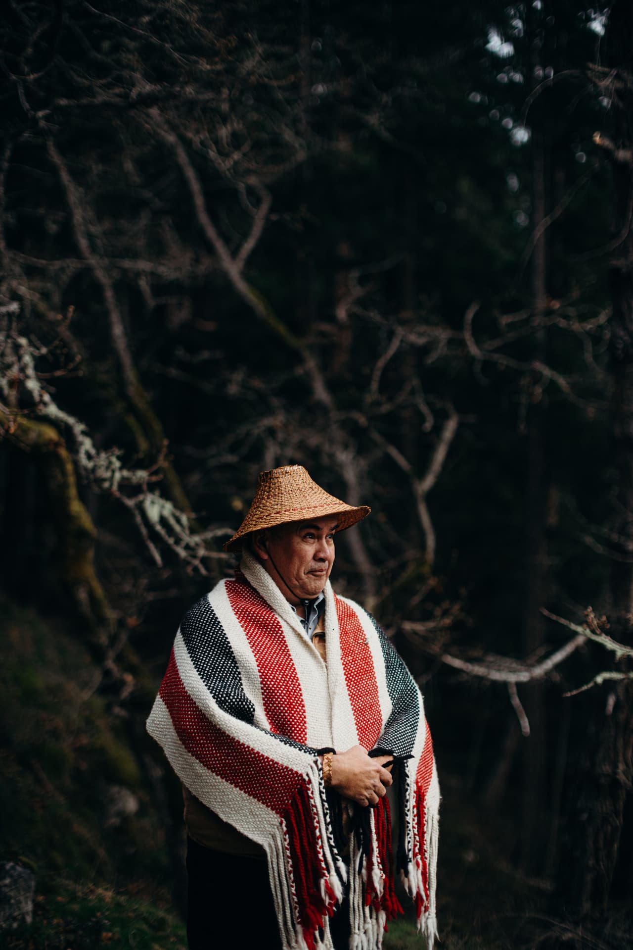 Man wearing woven hat and red-and-white striped blanket with fringe looks toward camera against dark forest background