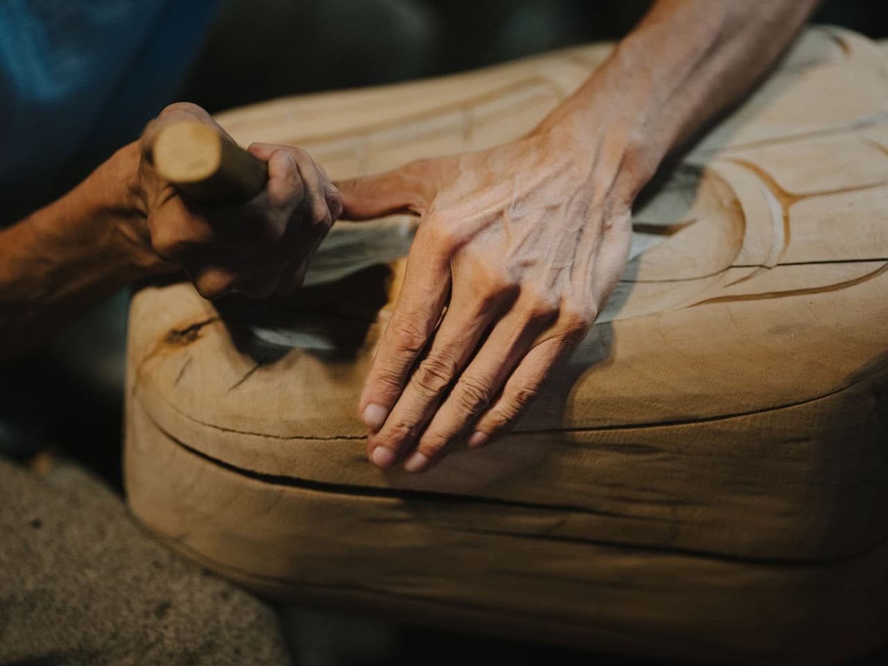 Weathered hands of a Port Hardy carver rest on pale wood in his workshop, light catching the texture of skin and grain