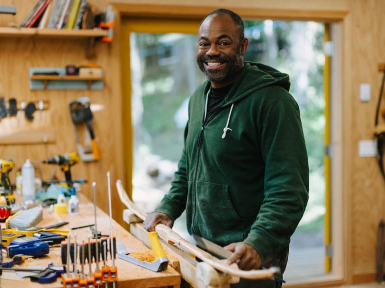 Christopher Applewhaite smiling at his workbench among drawknives and bow staves