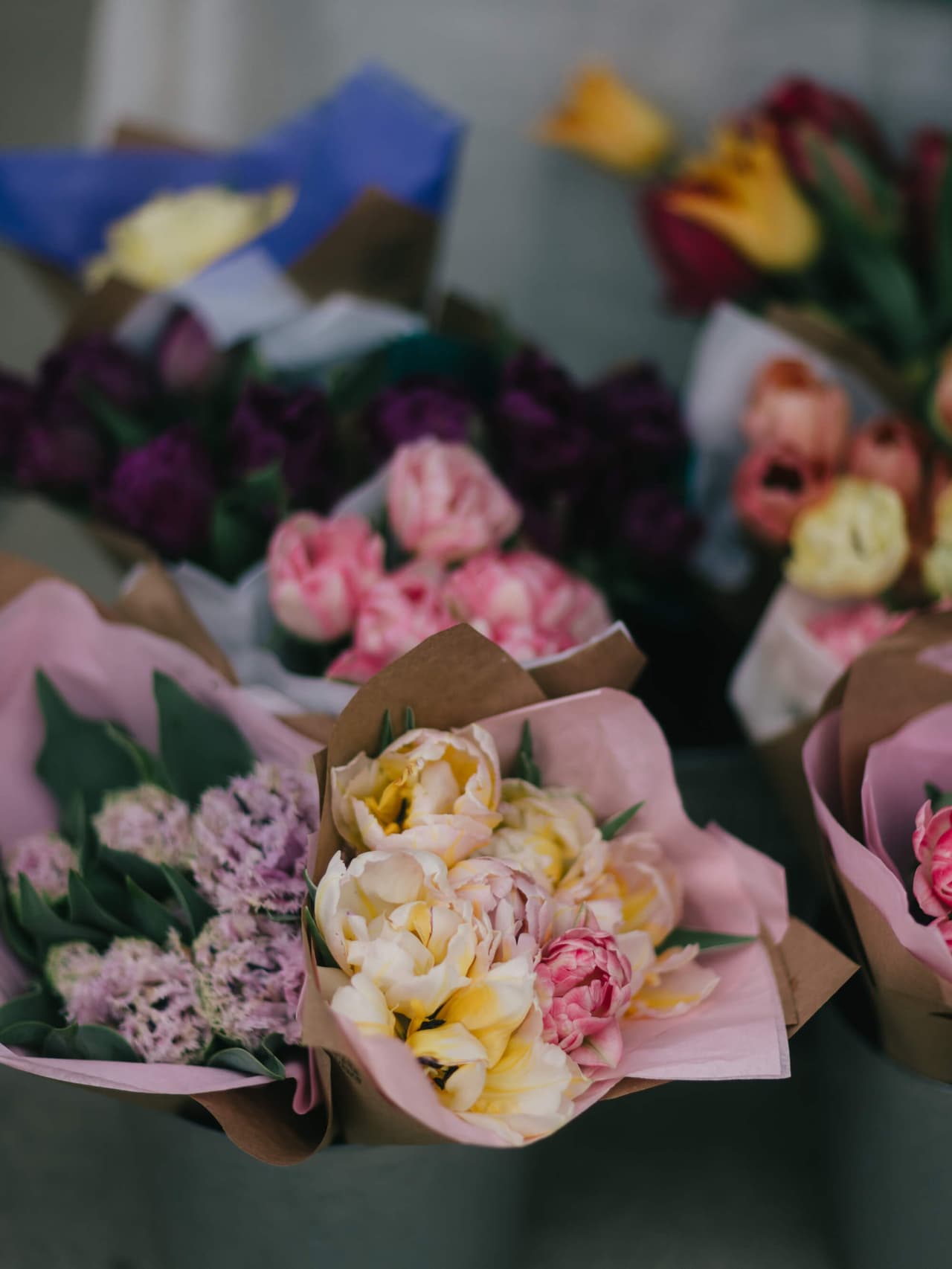 Close-up of pale yellow and pink peonies in paper wrapping, with blurred bouquets beyond at the market