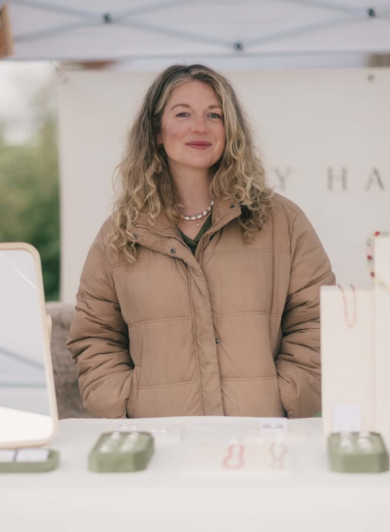 Woman in tan puffer jacket smiling at Saturday Market under white tent canopy