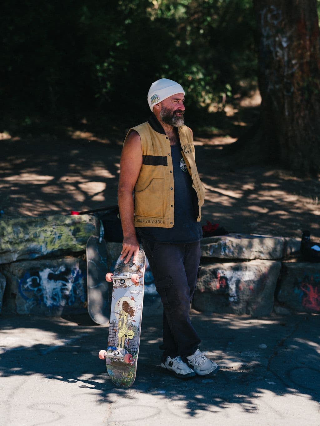 Bearded skateboarder holds decorated skateboard in dappled shade at Kanaka Road Skatepark