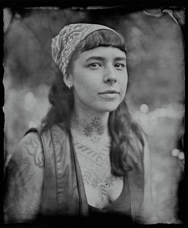Close profile of a person wearing a patterned headwrap, with decorative tattoos visible on neck and chest against dark background