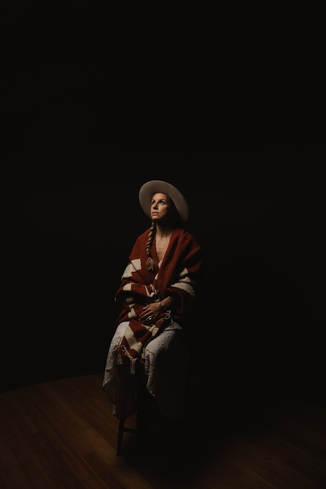 Woman in red and cream striped blanket and wide-brimmed hat gazes upward against black background, hands folded in lap