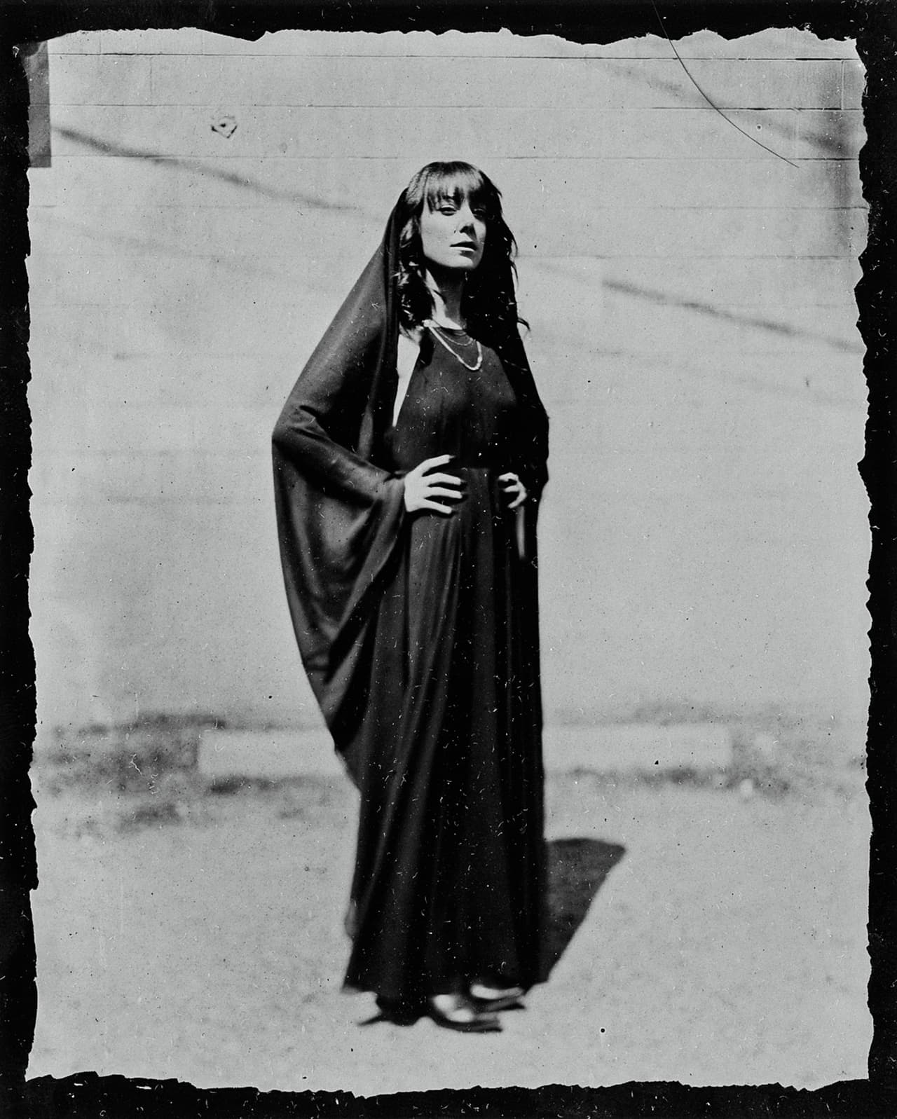 Woman in long dark dress with shawl and pearl necklace stands against textured wall, gazing upward in contemplative pose