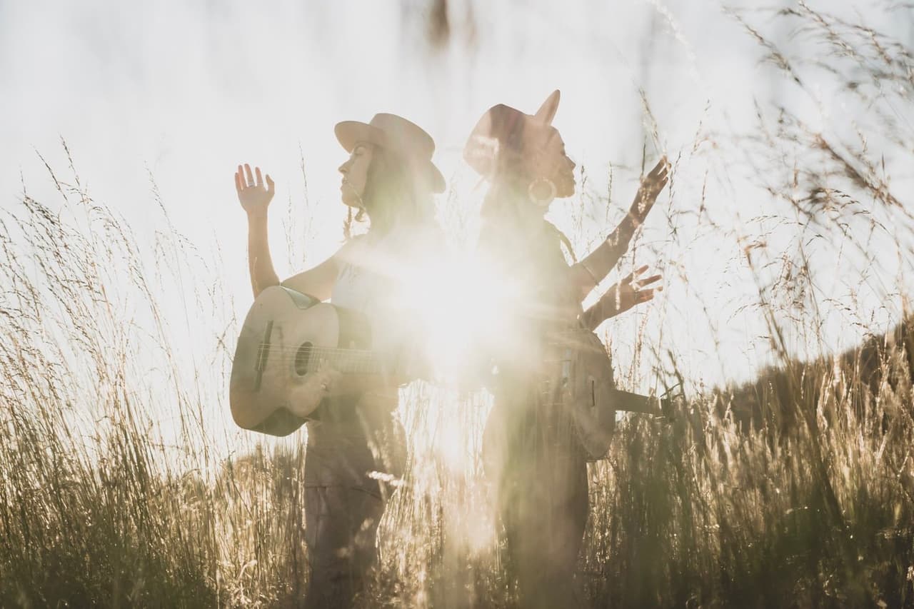 Two figures in cowboy hats stand in dried grass backlit by bright sunlight, faces obscured in silhouette and golden haze